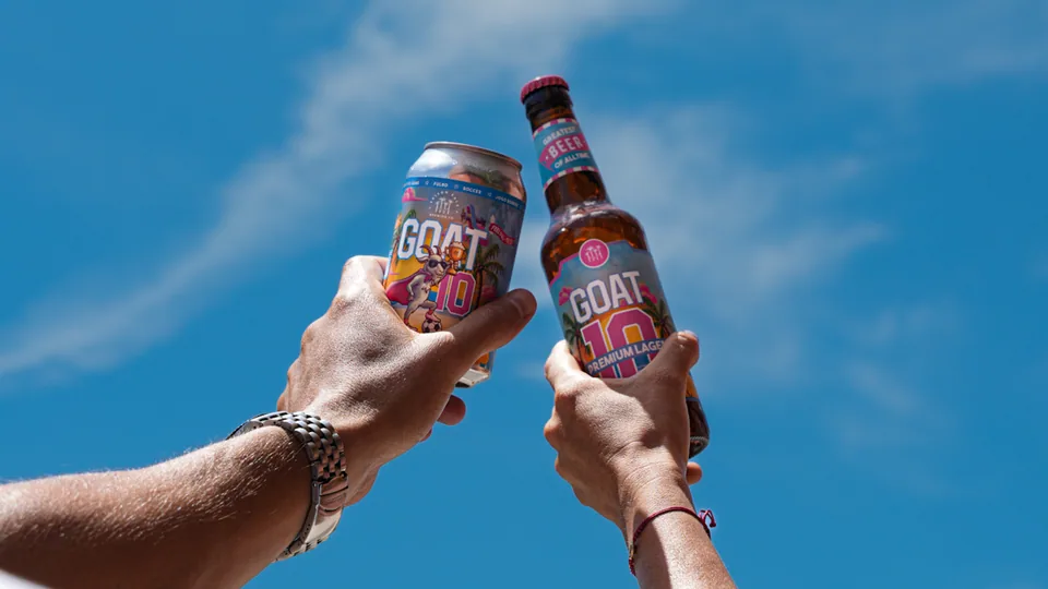A GOAT10 can and bottle raised together against a blue Miami sky.